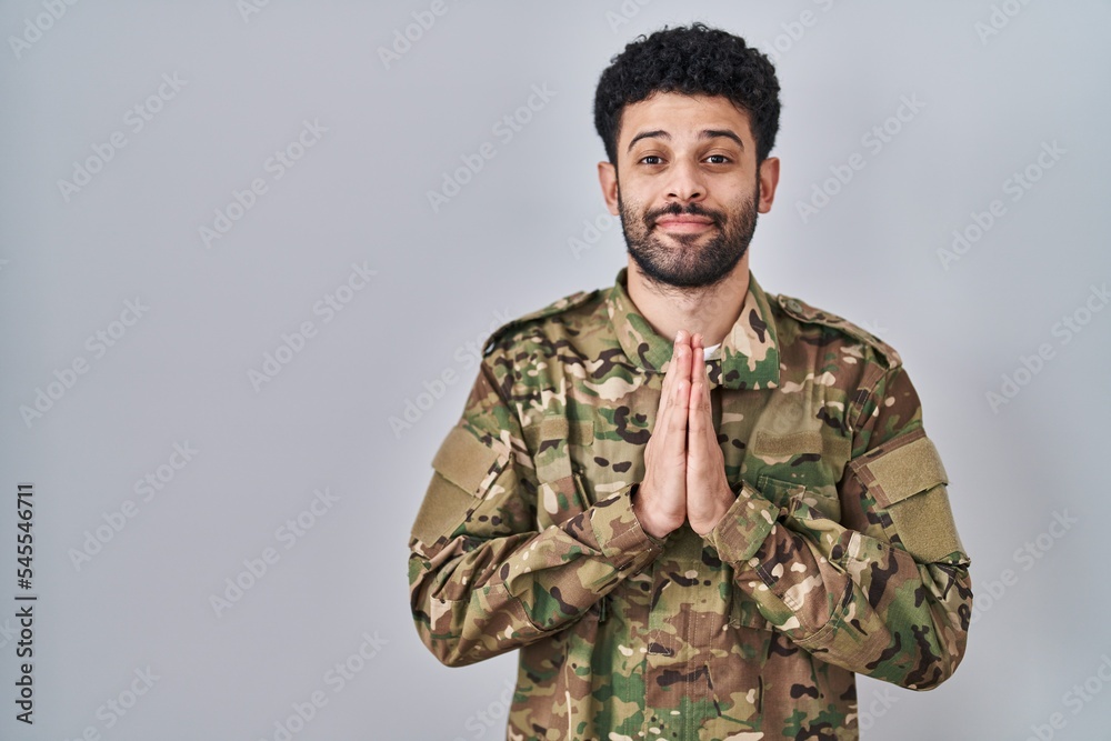 Arab man wearing camouflage army uniform praying with hands together asking for forgiveness smiling confident.