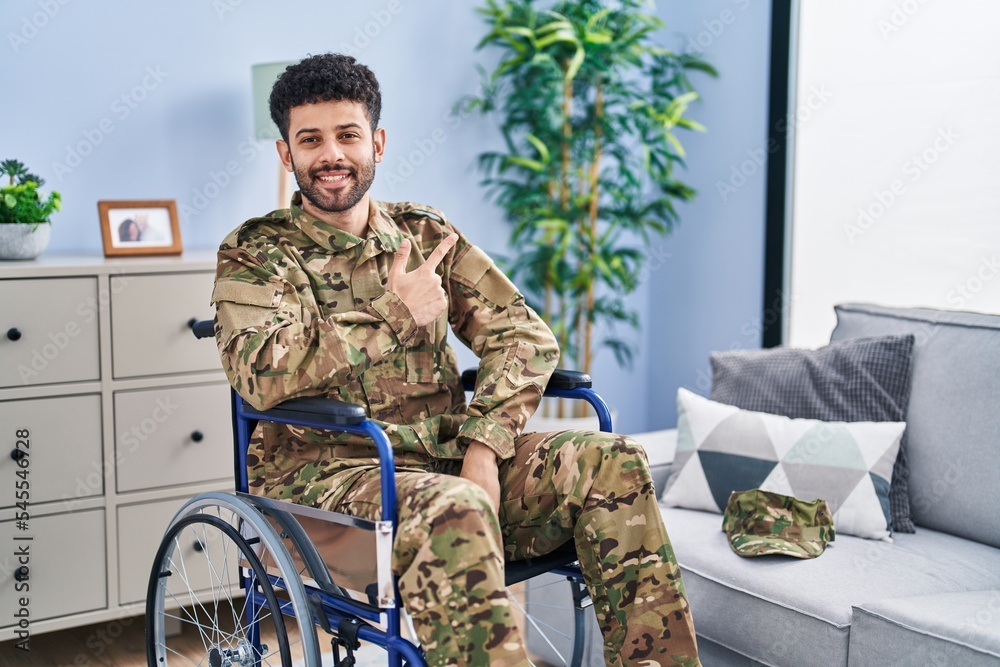 Arab man wearing camouflage army uniform sitting on wheelchair cheerful with a smile of face pointing with hand and finger up to the side with happy and natural expression on face