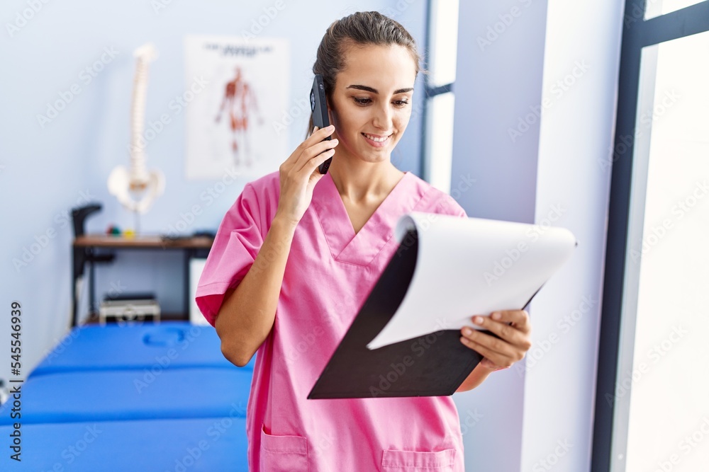Young hispanic woman wearing physiotherapist uniform talking on the smartphone at physiotherpy clinic Young hispanic woman wearing physiotherapist uniform talking on the smartphone at physiotherpy clinic