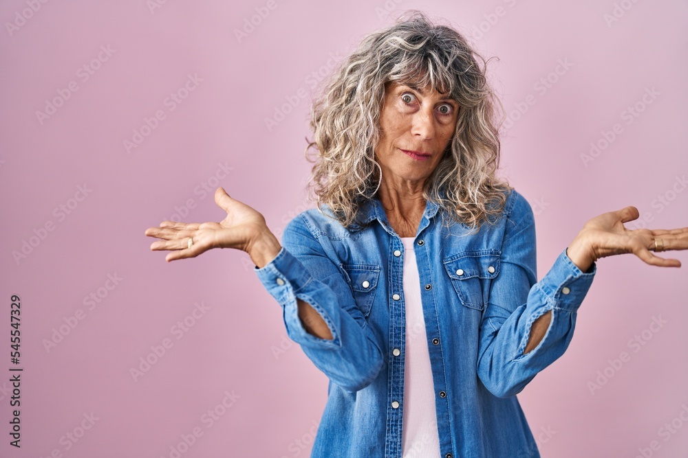 Middle age woman standing over pink background clueless and confused expression with arms and hands raised. doubt concept.