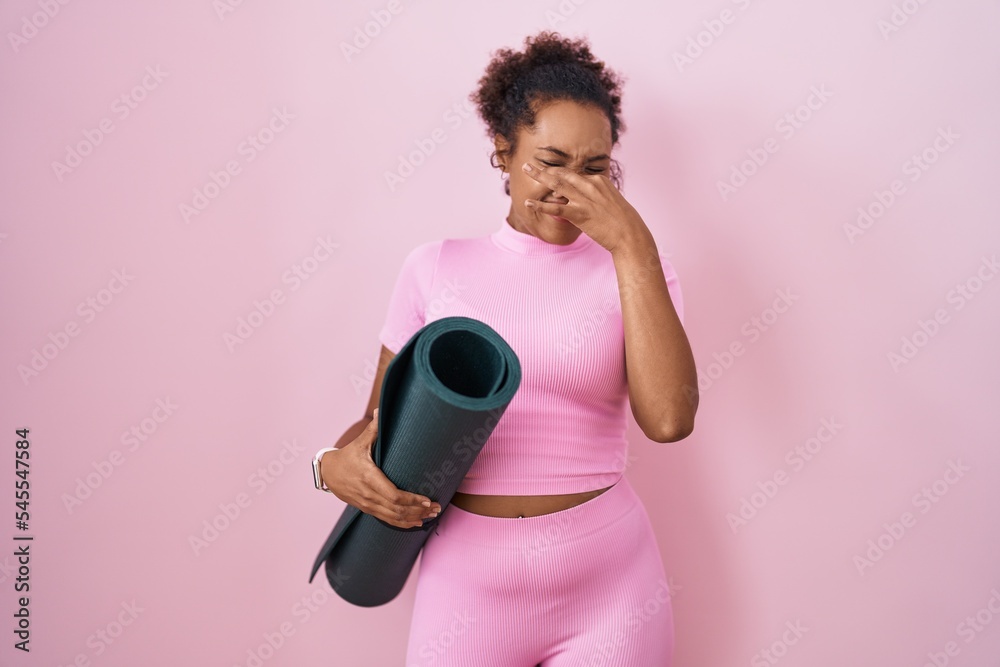 Young hispanic woman with curly hair holding yoga mat over pink background smelling something stinky and disgusting, intolerable smell, holding breath with fingers on nose. bad smell