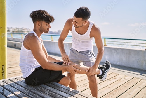 Two hispanic men sporty couple sitting on bench for leg injury at seaside