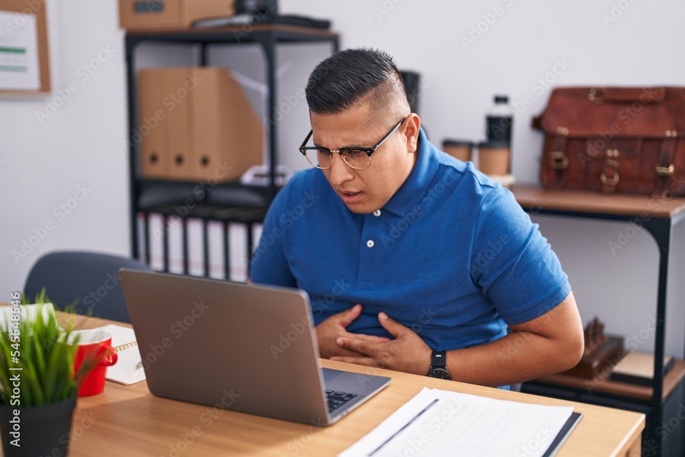 Young hispanic man working at the office with laptop with hand on stomach because indigestion, painful illness feeling unwell. ache concept. Young hispanic man working at the office with laptop with hand on stomach because indigestion, painful illness feeling unwell. ache concept.