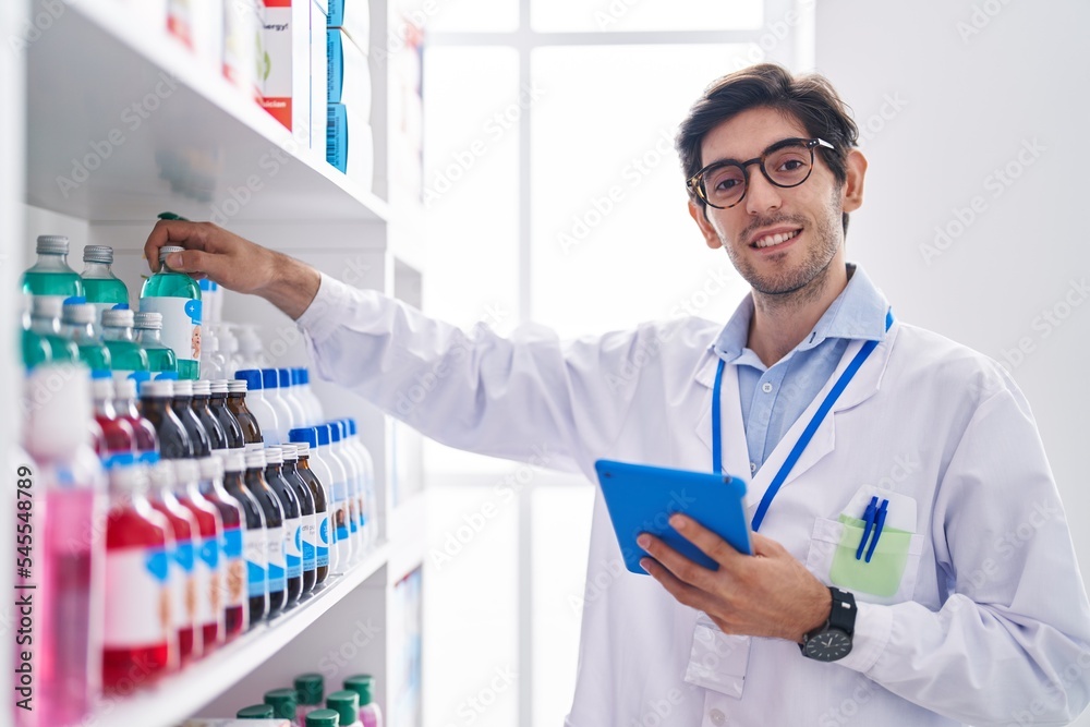Young hispanic man pharmacist using touchpad holding medicine at pharmacy
