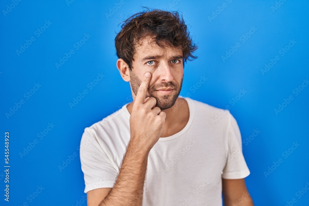 Hispanic young man standing over blue background pointing to the eye watching you gesture, suspicious expression Hispanic young man standing over blue background pointing to the eye watching you gesture, suspicious expression