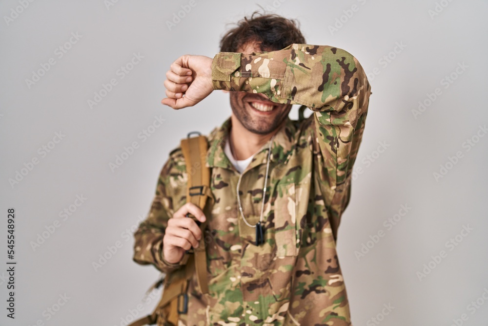 Hispanic young man wearing camouflage army uniform smiling cheerful playing peek a boo with hands showing face. surprised and exited