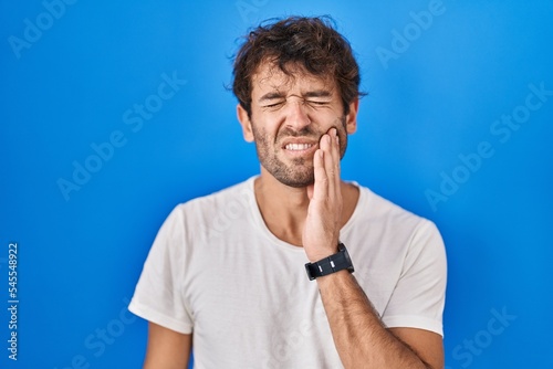 Hispanic young man standing over blue background touching mouth with hand with painful expression because of toothache or dental illness on teeth. dentist