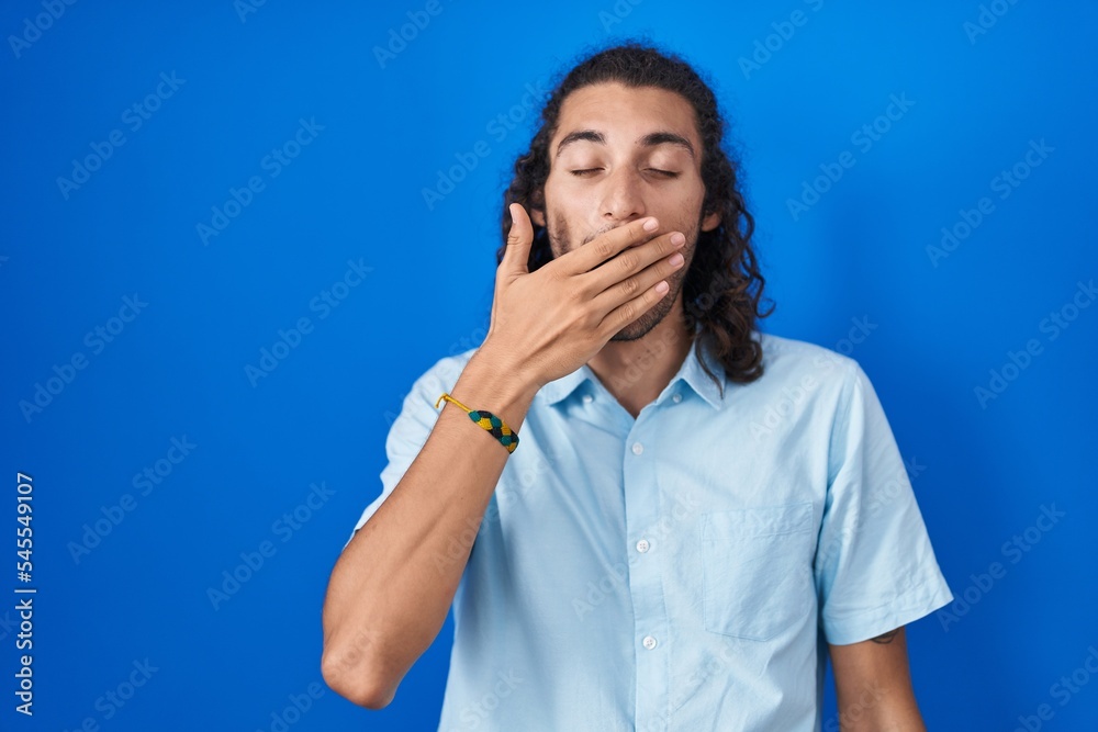 Young hispanic man standing over blue background bored yawning tired covering mouth with hand. restless and sleepiness. Young hispanic man standing over blue background bored yawning tired covering mouth with hand. restless and sleepiness.