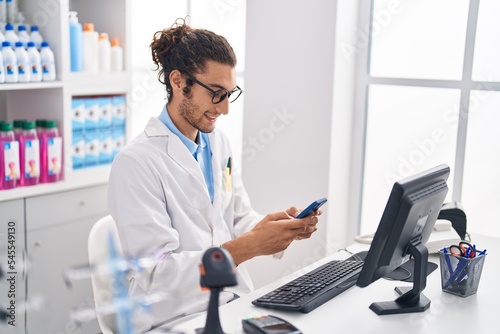 Young hispanic man pharmacist using smartphone working at pharmacy photo