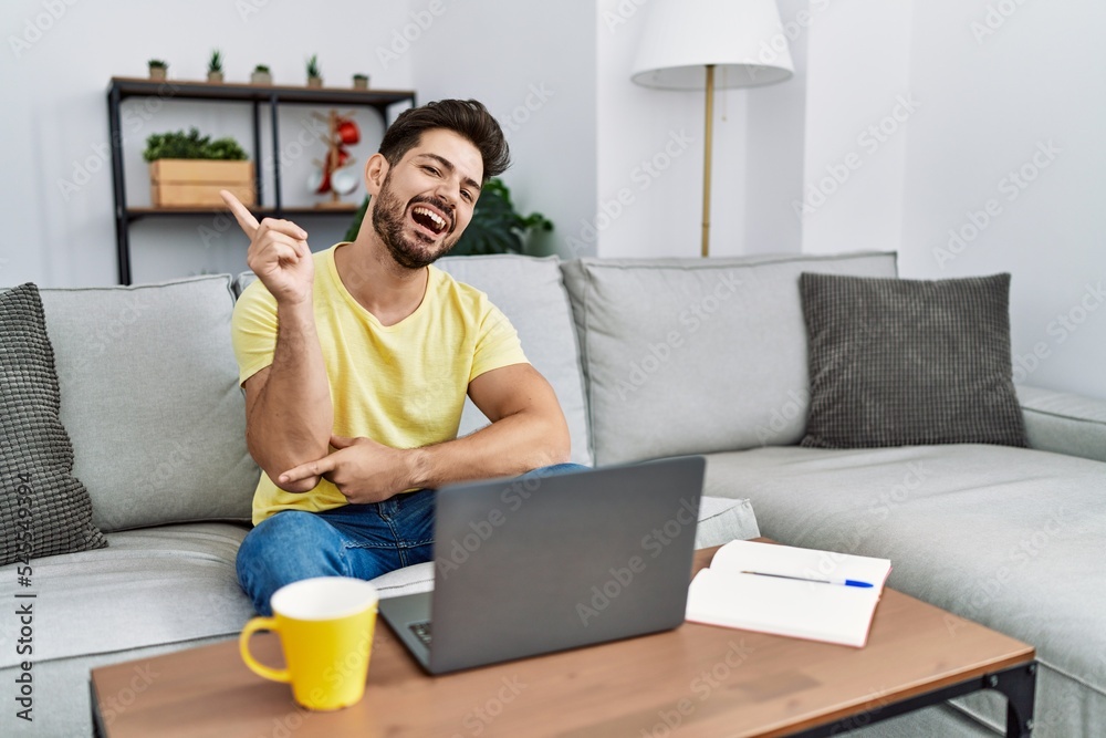 Young man with beard using laptop at home with a big smile on face, pointing with hand and finger to the side looking at the camera. Young man with beard using laptop at home with a big smile on face, pointing with hand and finger to the side looking at the camera.