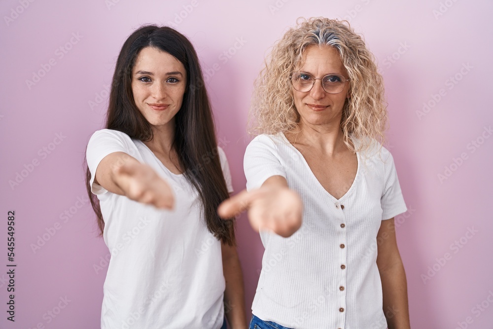 Mother and daughter standing together over pink background smiling cheerful offering palm hand giving assistance and acceptance. Mother and daughter standing together over pink background smiling cheerful offering palm hand giving assistance and acceptance.