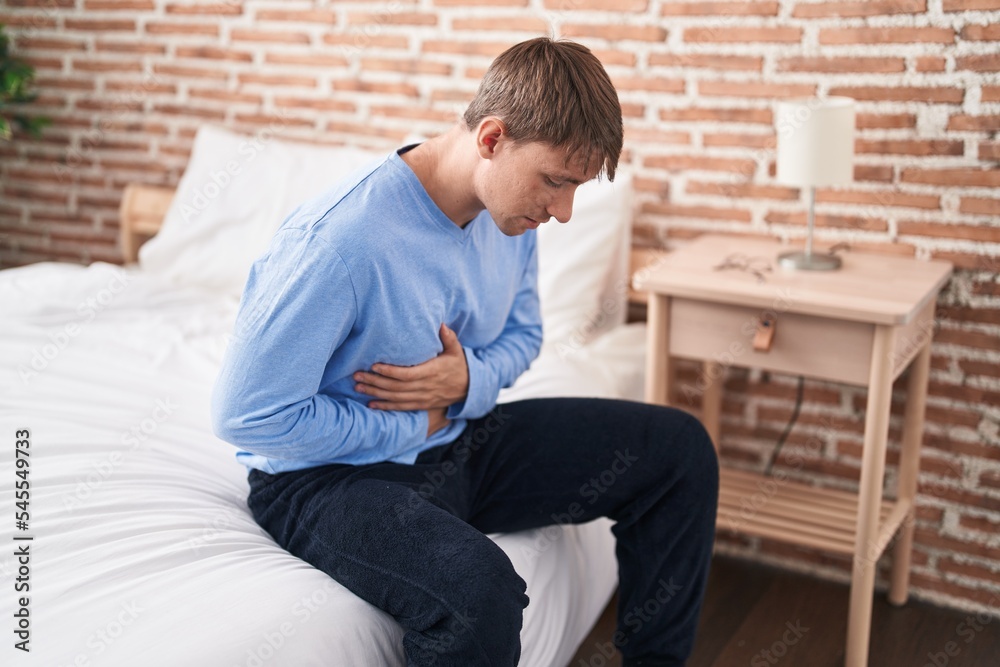 Young caucasian man suffering for stomach ache sitting on bed at bedroom