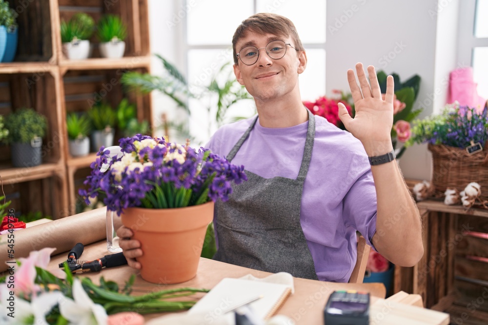 Caucasian blond man working at florist shop waiving saying hello happy and smiling, friendly welcome gesture