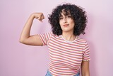 Young middle east woman standing over pink background strong person showing arm muscle, confident and proud of power