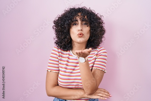Young middle east woman standing over pink background looking at the camera blowing a kiss with hand on air being lovely and sexy. love expression.