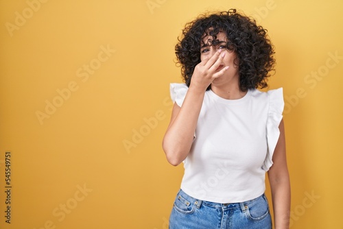 Young middle east woman standing over yellow background smelling something stinky and disgusting, intolerable smell, holding breath with fingers on nose. bad smell