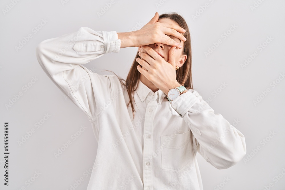 Young caucasian woman standing over isolated background covering eyes and mouth with hands, surprised and shocked. hiding emotion Young caucasian woman standing over isolated background covering eyes and mouth with hands, surprised and shocked. hiding emotion