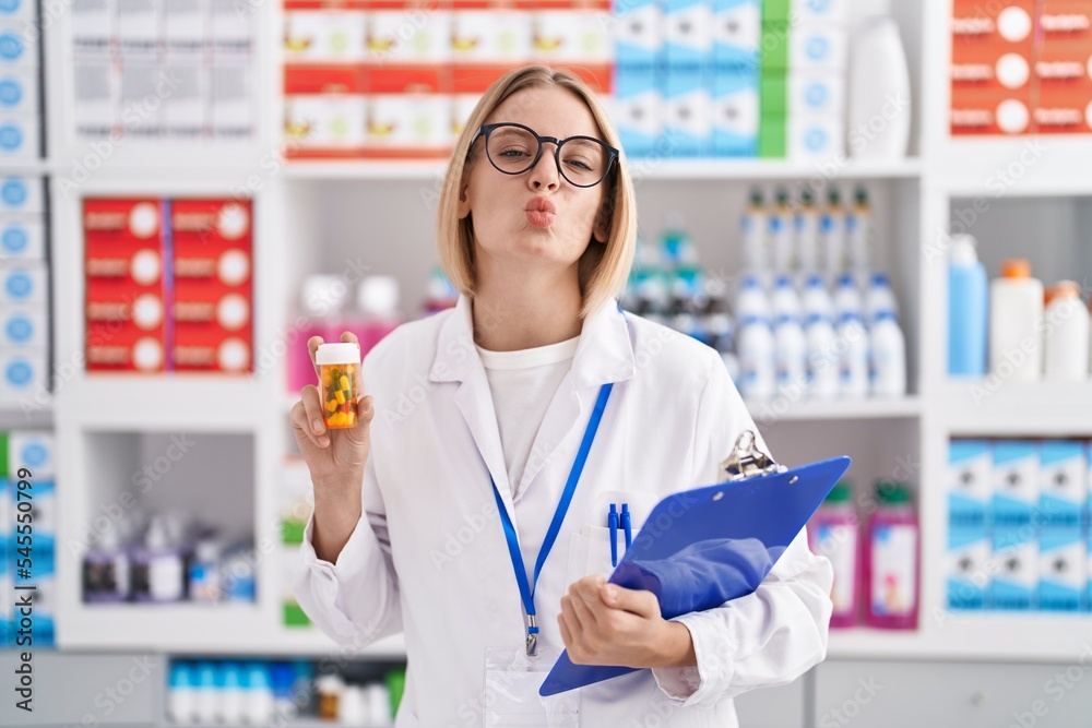 Young caucasian woman working at pharmacy drugstore holding pills looking at the camera blowing a kiss being lovely and sexy. love expression.