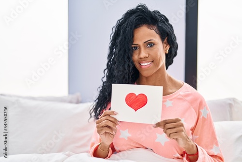Middle age hispanic woman sitting on the bed at home looking positive and happy standing and smiling with a confident smile showing teeth