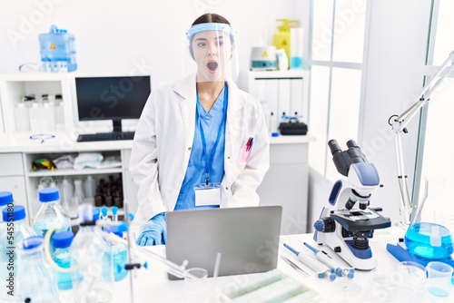 Young hispanic woman wearing scientist uniform scared and amazed with open mouth for surprise  disbelief face
