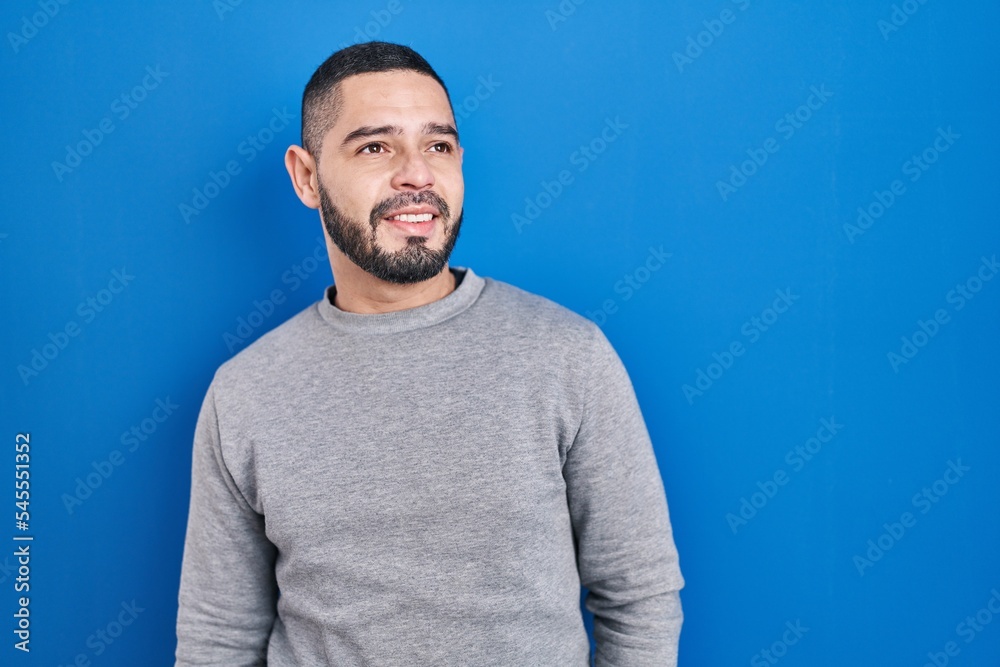 Hispanic man standing over blue background looking away to side with smile on face, natural expression. laughing confident. Hispanic man standing over blue background looking away to side with smile on face, natural expression. laughing confident.