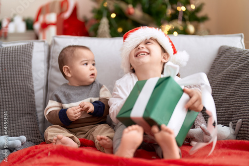 Brother and sister opening gift sitting on sofa by christmas tree at home