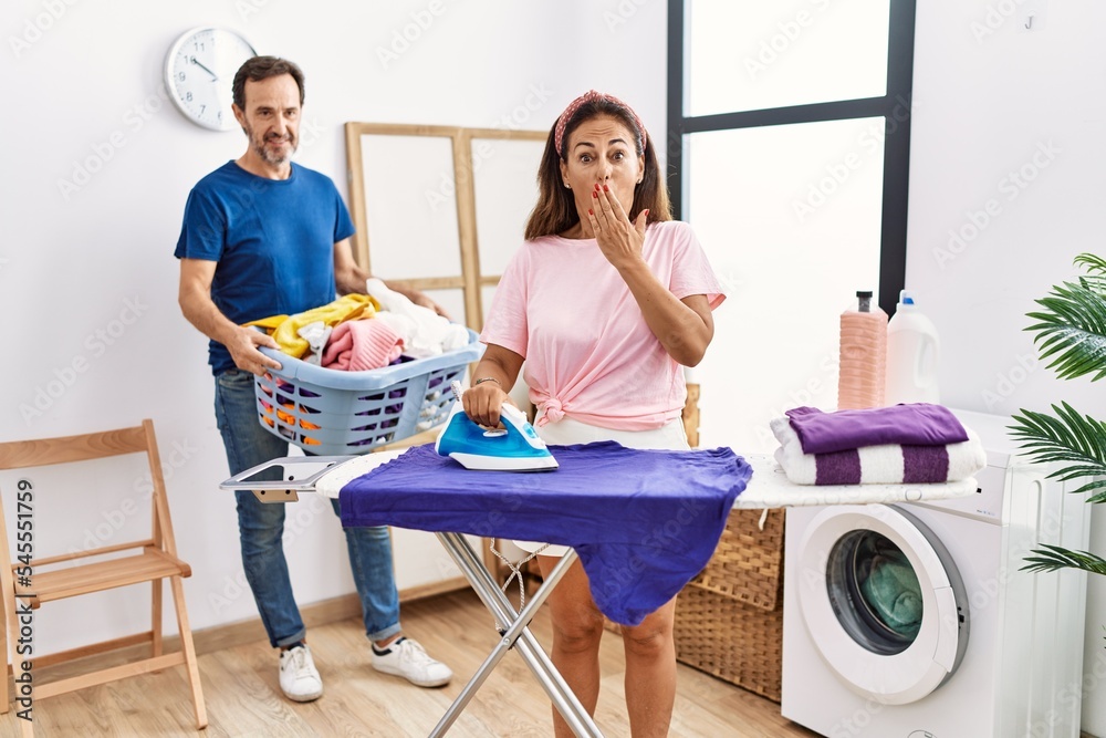 Obraz premium Middle age couple ironing clothes at home covering mouth with hand, shocked and afraid for mistake. surprised expression
