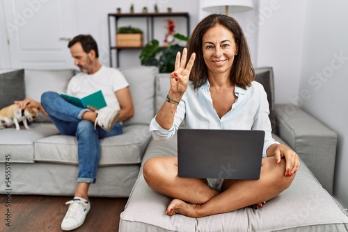 Hispanic middle age couple at home, woman using laptop showing and pointing up with fingers number three while smiling confident and happy.