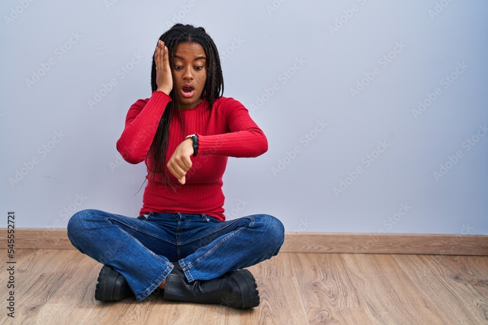 Young african american with braids sitting on the floor at home looking at the watch time worried, afraid of getting late
