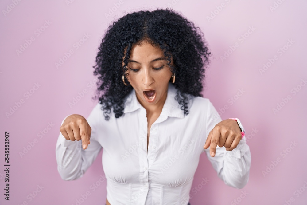 Hispanic woman with curly hair standing over pink background pointing down with fingers showing advertisement, surprised face and open mouth