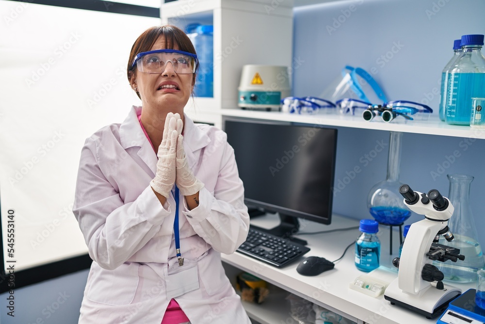 Young brunette woman working at scientist laboratory begging and praying with hands together with hope expression on face very emotional and worried. begging. Young brunette woman working at scientist laboratory begging and praying with hands together with hope expression on face very emotional and worried. begging.