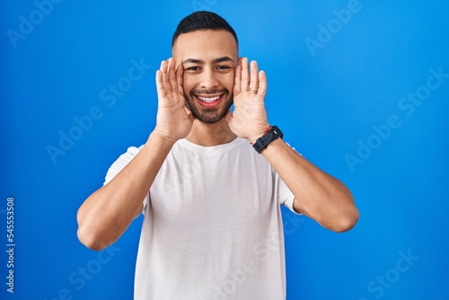 Young hispanic man standing over blue background smiling cheerful playing peek a boo with hands showing face. surprised and exited