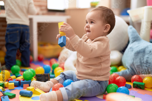 Adorable toddler playing with wooden construction blocks sitting on floor at kindergarten