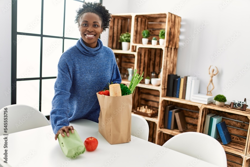 African american woman smiling confident holding food of groceries bag at home African american woman smiling confident holding food of groceries bag at home