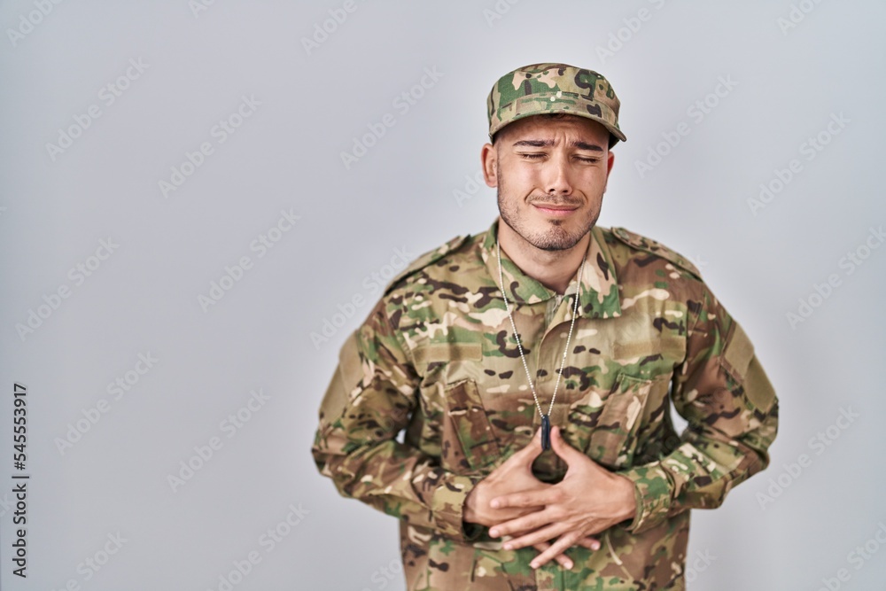 Young hispanic man wearing camouflage army uniform with hand on stomach because nausea, painful disease feeling unwell. ache concept. Young hispanic man wearing camouflage army uniform with hand on stomach because nausea, painful disease feeling unwell. ache concept.