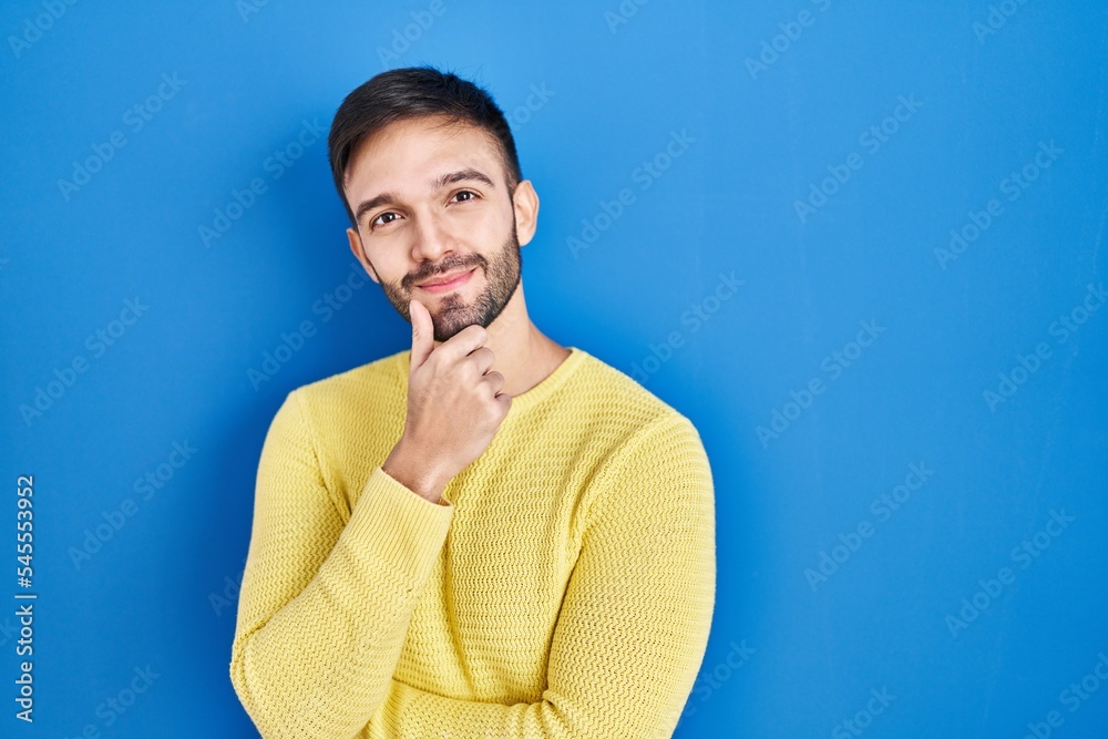 Hispanic man standing over blue background looking confident at the camera smiling with crossed arms and hand raised on chin. thinking positive. Hispanic man standing over blue background looking confident at the camera smiling with crossed arms and hand raised on chin. thinking positive.