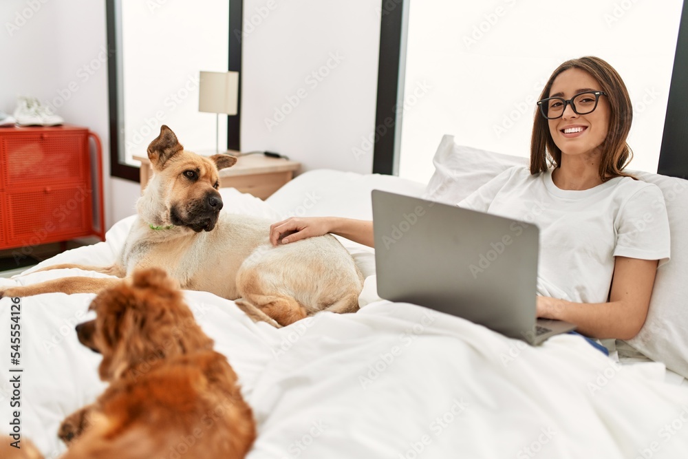 Obraz premium Young hispanic woman using laptop sitting on bed with dogs at bedroom