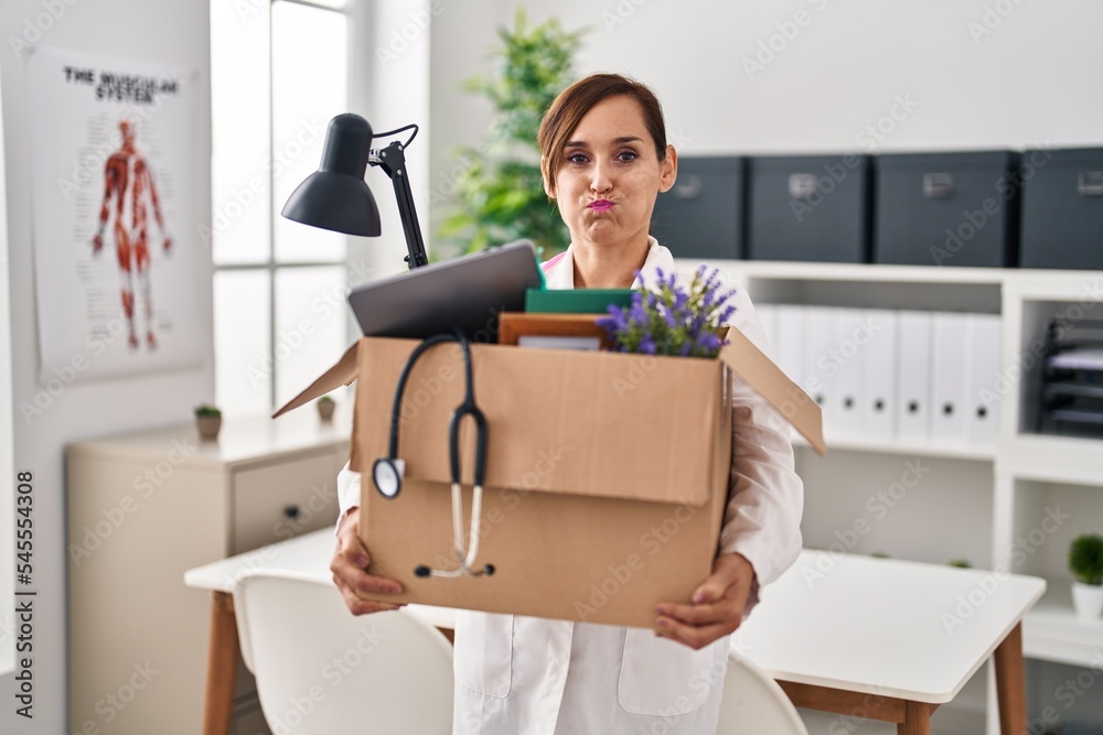 Middle age doctor woman holding cardboard box with items at the clinic puffing cheeks with funny face. mouth inflated with air, catching air. Middle age doctor woman holding cardboard box with items at the clinic puffing cheeks with funny face. mouth inflated with air, catching air.