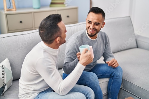 Two men couple drinking coffee sitting on sofa at home