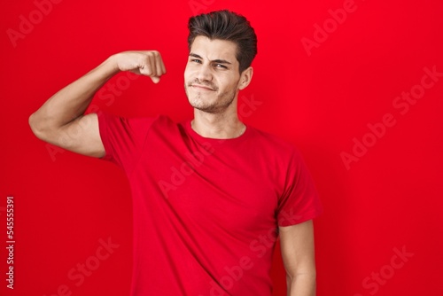 Young hispanic man standing over red background strong person showing arm muscle, confident and proud of power © Krakenimages.com