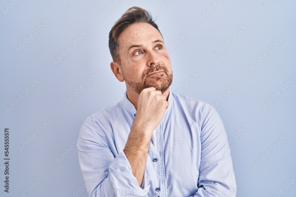 Middle age caucasian man standing over blue background with hand on chin thinking about question, pensive expression. smiling with thoughtful face. doubt concept.