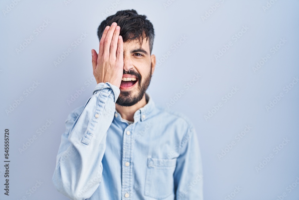 Young hispanic man with beard standing over blue background covering one eye with hand, confident smile on face and surprise emotion. Young hispanic man with beard standing over blue background covering one eye with hand, confident smile on face and surprise emotion.