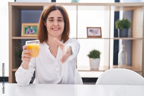 Brunette woman drinking glass of orange juice smiling friendly offering handshake as greeting and welcoming. successful business.