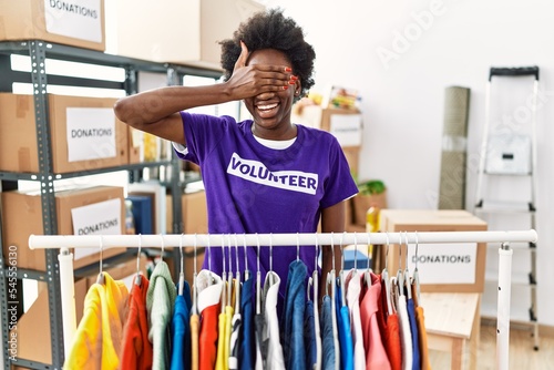African young woman wearing volunteer t shirt at donations stand smiling and laughing with hand on face covering eyes for surprise. blind concept.