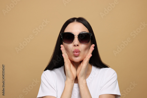 Beautiful young woman in stylish sunglasses blowing kiss on beige background