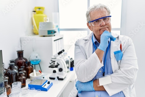 Middle age grey-haired man wearing scientist uniform with arms crossed gesture at laboratory