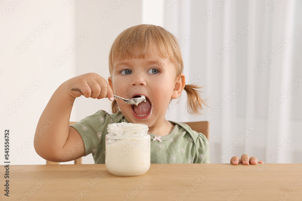 Cute little child eating tasty yogurt with spoon at wooden table indoors Cute little child eating tasty yogurt with spoon at wooden table indoors