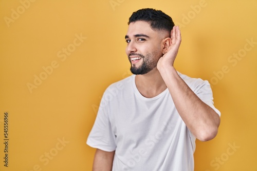 Young handsome man wearing casual t shirt over yellow background smiling with hand over ear listening an hearing to rumor or gossip. deafness concept.