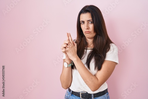 Young brunette woman standing over pink background holding symbolic gun with hand gesture  playing killing shooting weapons  angry face
