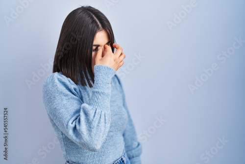 Young hispanic woman standing over blue background tired rubbing nose and eyes feeling fatigue and headache. stress and frustration concept. © Krakenimages.com